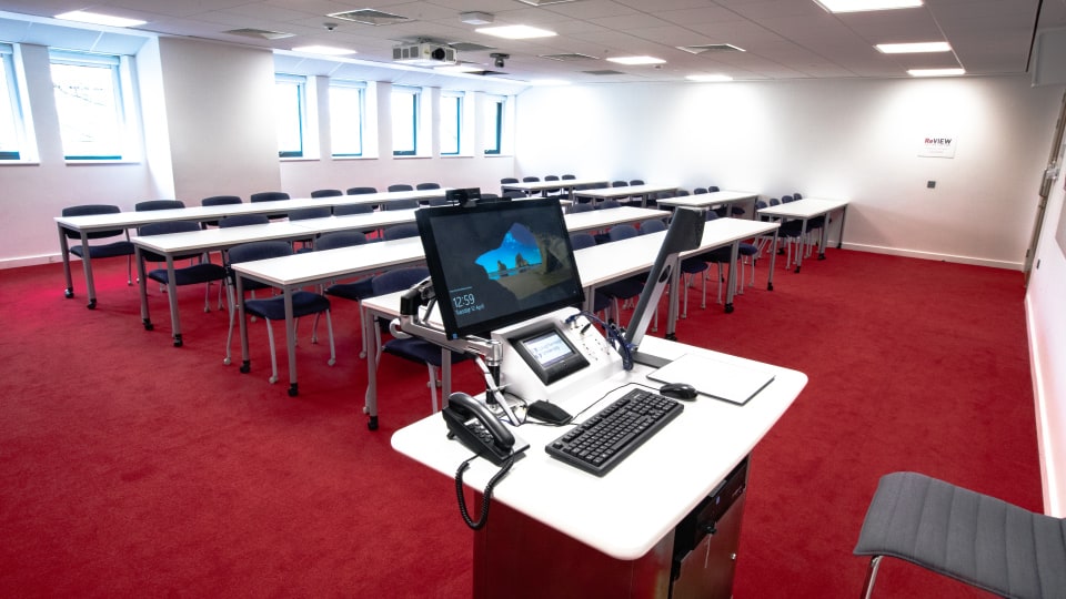 David Davies DAV1100 - Teaching room with red carpet, rows of tables and chairs, and a lectern with computer and phone. Bright windows.
