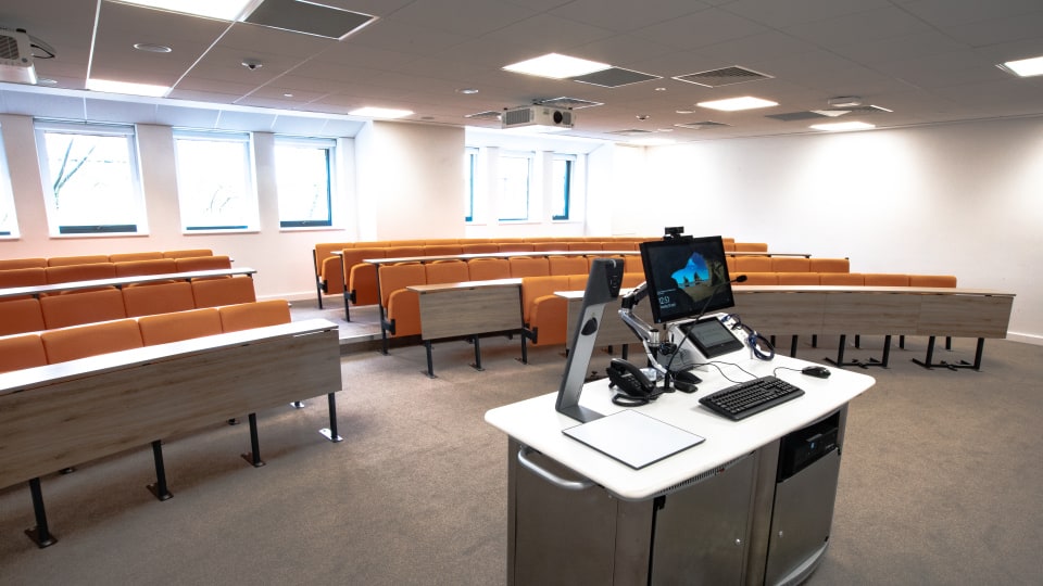 David Davies DAV1101 - Lecture theatre with tiered curved orange seating, wooden desks, and a central teaching podium equipped with a computer and projector.