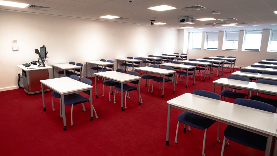 David Davies DAV1102 - Classroom with red carpet, white walls, and rows of white tables with blue chairs. Instructor's desk with computer at front.