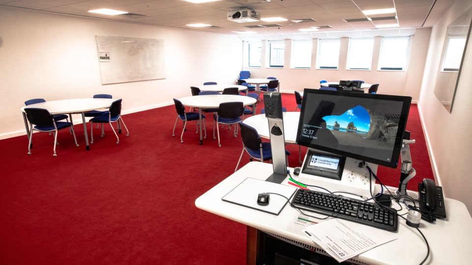 David Davies DAV1103 - Teaching room with red carpet, circular tables, blue chairs, whiteboard, and instructor's desk with computer and projector.