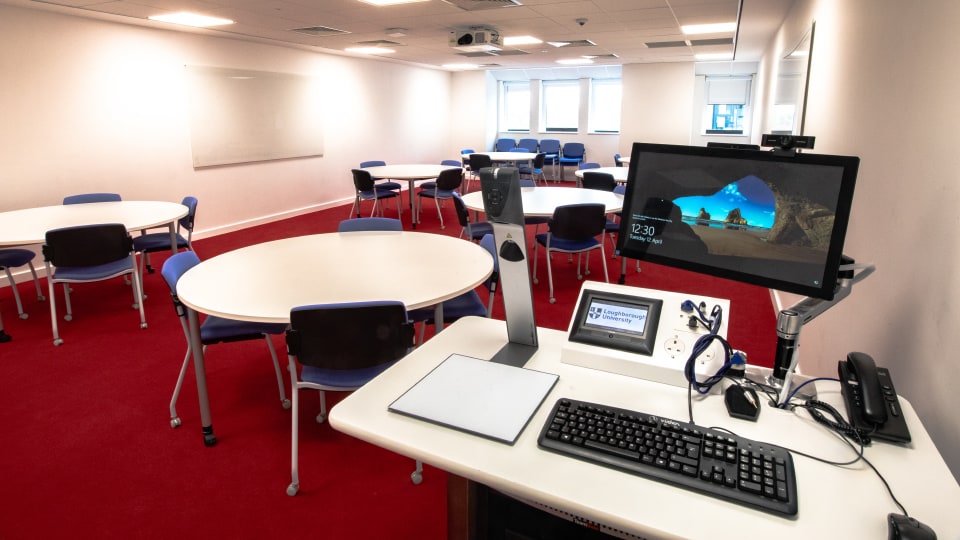 David Davies DAV1104 - Teaching room with red carpet, round tables, blue chairs, and a presentation desk featuring a computer and control panel.
