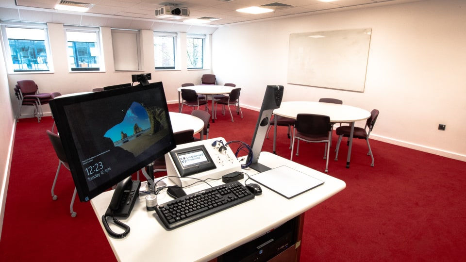 David Davies DAV1105 - Teaching room with red carpet, round tables, chairs, whiteboard, and a podium featuring a computer and control panel.