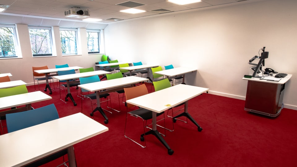 David Davies DAV1106 - Classroom with red carpet, colourful chairs, white desks, projector, and instructor's podium with computer near windows.