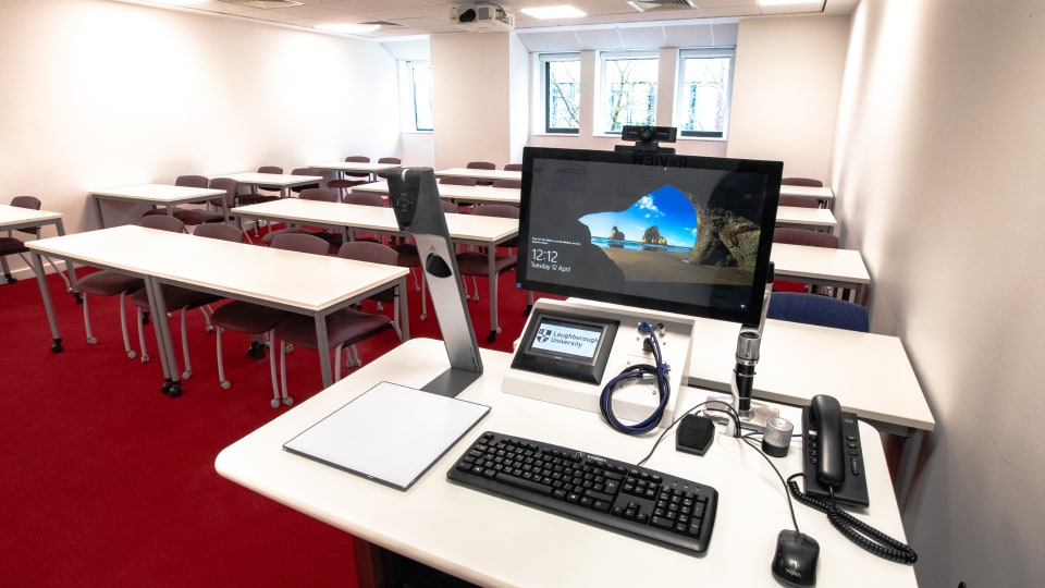 David Davies DAV1107 - Teaching room with red carpet, rows of desks and chairs. Front desk has computer, keyboard, phone, and cables.