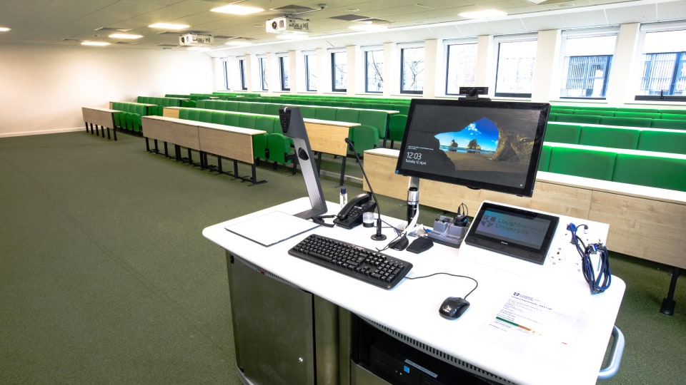 David Davies DAV1108 - Lecture theatre with green tiered seating, large windows, and a teaching podium equipped with computer, microphone, and control panel.