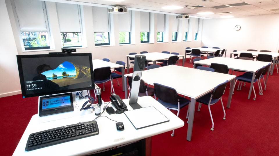 David Davies DAV1109 - Teaching room with red carpet, white tables, blue chairs, and a lectern with computer, keyboard, and phone. Windows line the wall.