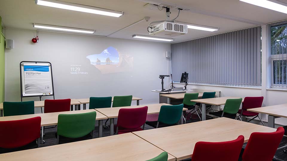 Teaching Room E007 - Classroom with colourful chairs, desks, projector, flipchart pad, and blinds. Projected screen displays time and date.