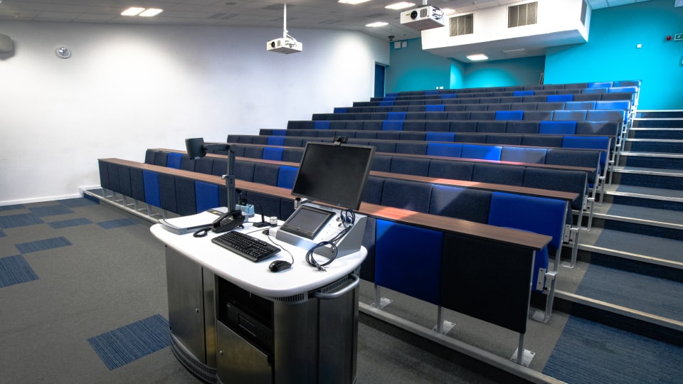 Edward Herbert Lecture EHB001 -  theatre with tiered seating, blue and gray chairs, and a podium equipped with computer and projector.