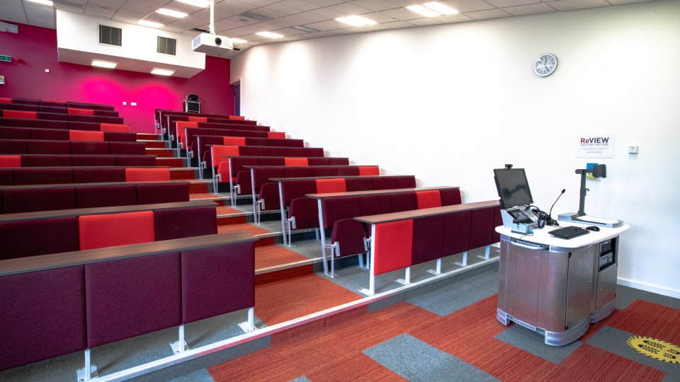Edward Herbert EHB002 - Lecture hall with tiered red seating, a whiteboard, and a podium with computer equipment. Bright lighting and clock.
