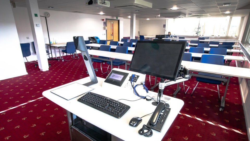 Edward Herbert EHB205 - Teaching room with red carpet, blue chairs, white tables, and a lectern featuring a computer and keyboard.