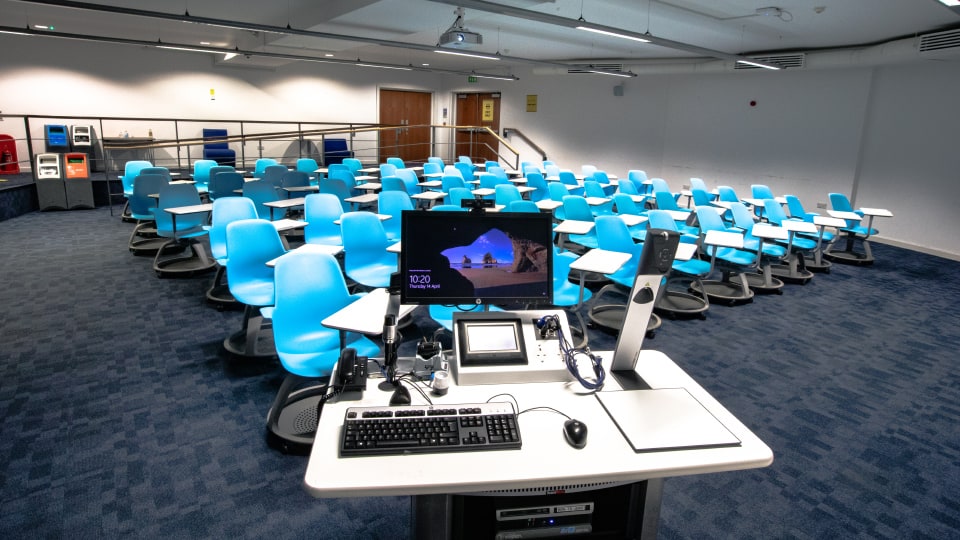 John Beckwith JB021 - Flat space with blue swivel chairs, desks, and a central teaching podium equipped with computer, microphone, and control panel.