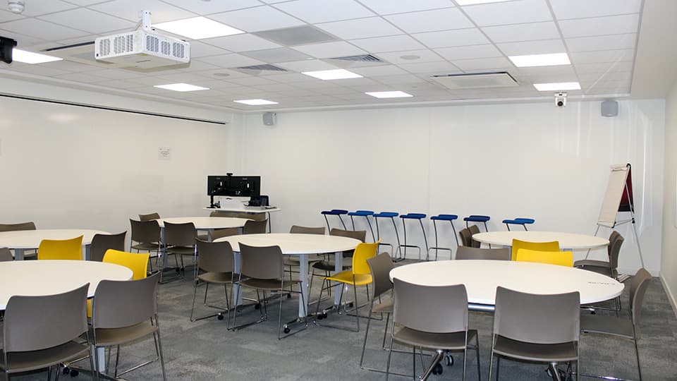 Medium-small white walled teaching space with round white desks and grey carpet.