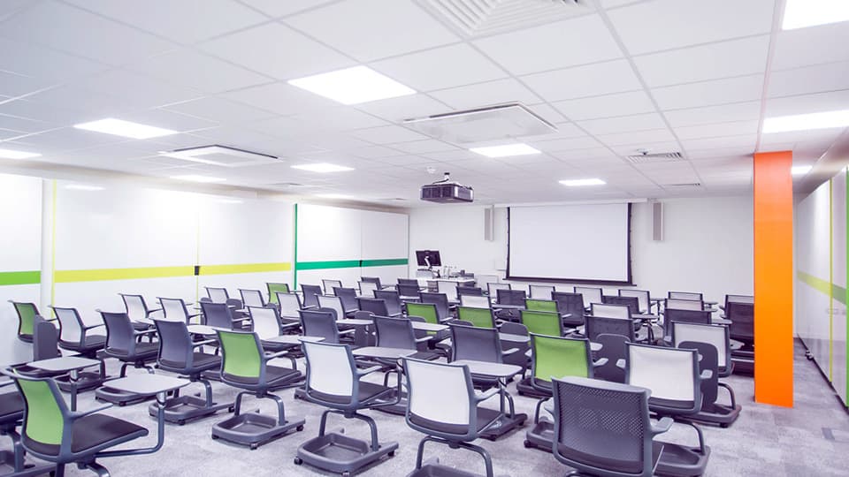 Large white teaching room with splashes of colour and individual black seat-desks in rows.