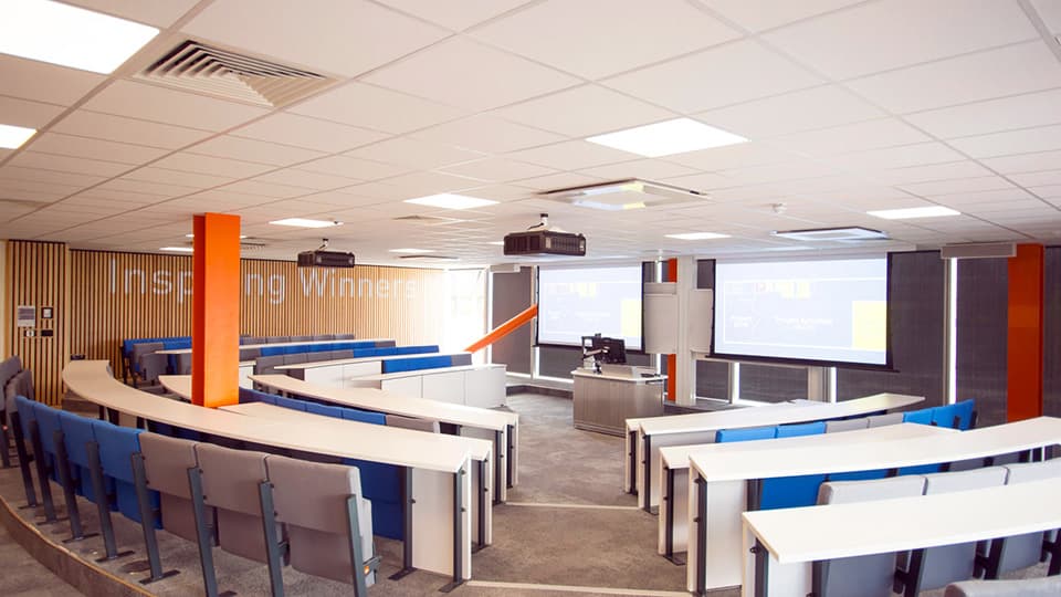 Semi-circular teaching space with white and blue seating at different levels, some pale strip wood panelling and two screens.