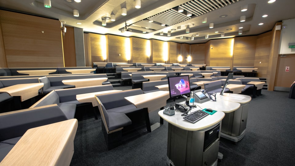 Design School LDS017 - Modern lecture theatre with tiered seating, wooden desks, and a central podium equipped with a computer and microphone.