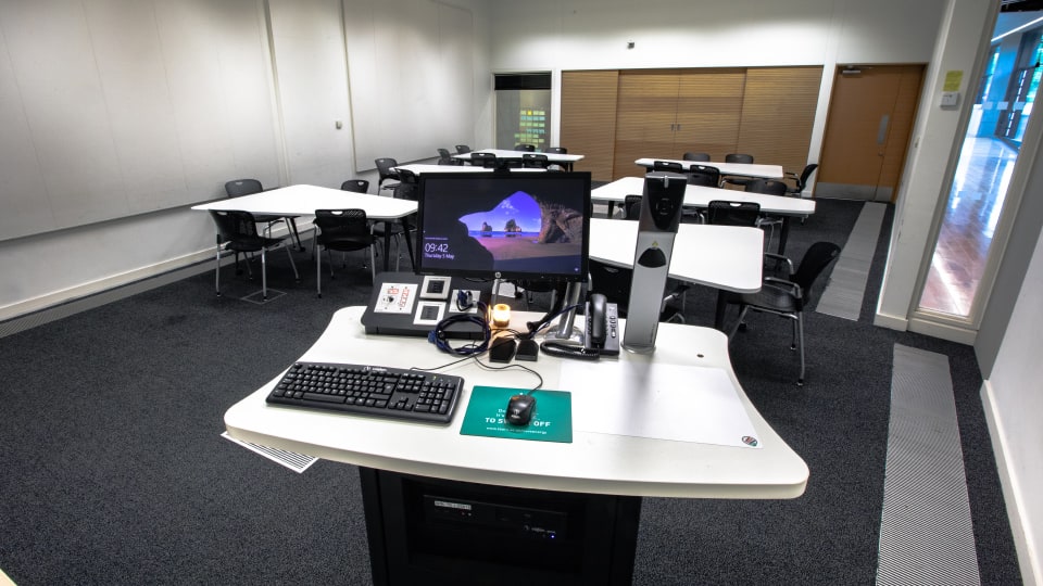 Design School LDS018 - Teaching room with tables, chairs, and a lectern featuring a computer, keyboard, and control panel.