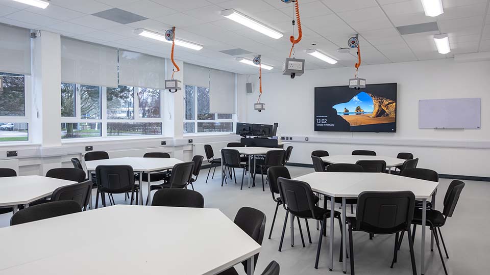 Teaching Room MHL011 - Modern teaching room with round tables, chairs, large screen, and ceiling-mounted projectors. Bright windows and whiteboard visible.