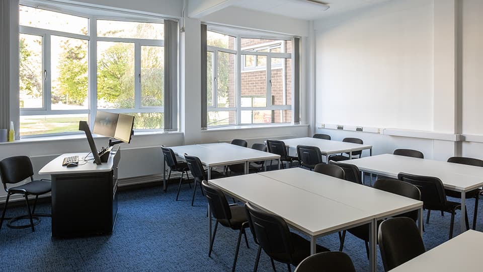 Medium-sized lecture room in Martin Hall with white desks set in groups of four, with white walls and blue carpet.