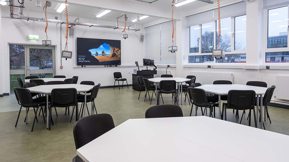 Teaching Room MHL013 - Modern teaching room with round tables, chairs, large screen, and windows. Bright lighting and visible outdoor greenery.