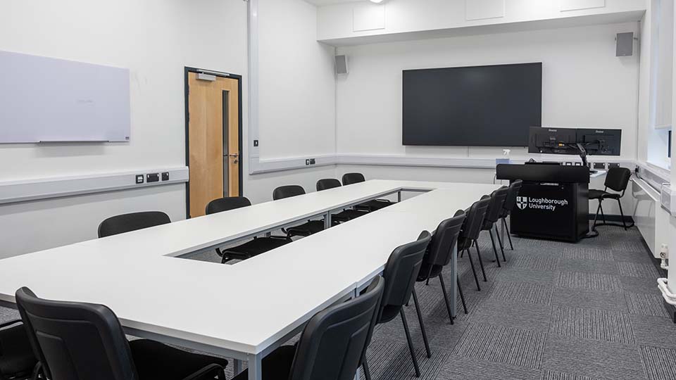 Teaching Room MHL015 - Teaching room with U-shaped table, black chairs, large screen, and lectern. Whiteboard on wall. Loughborough University branding.