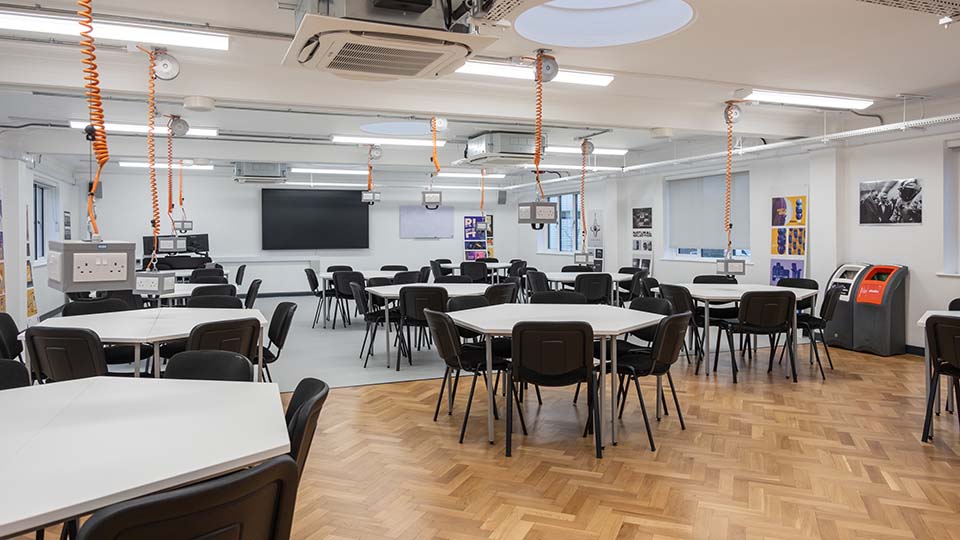 Teaching Room MHL111 - Spacious teaching room with hexagonal tables, black chairs, large screen, and wall displays. Bright lighting and parquet flooring.