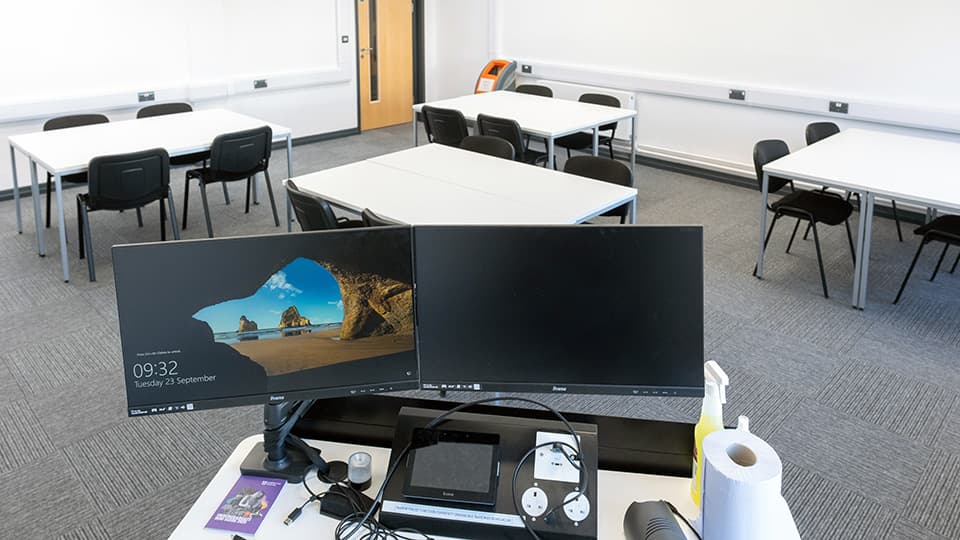 Medium-sized lecture room in Martin Hall with white desks set in groups of four, with white walls and grey carpet, viewed from the lecturer's area.