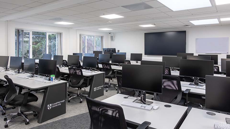 Teaching Room MHL117 - Computer lab with rows of desks, monitors, and chairs. Large screen and whiteboard at the front.