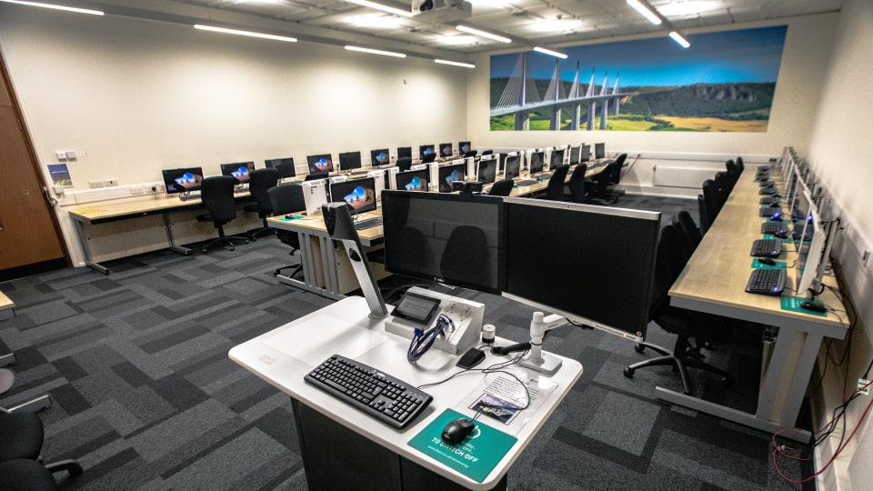 Frank Gibb RT026 - Computer lab with rows of desks, computers, and chairs. Instructor's station in foreground. Large mural of a bridge on the wall.