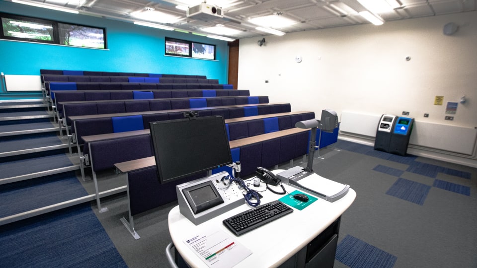 Frank Gibb RT033 - Lecture theatre with tiered seating, blue accents, and a teaching podium equipped with a computer, keyboard, and control panel.
