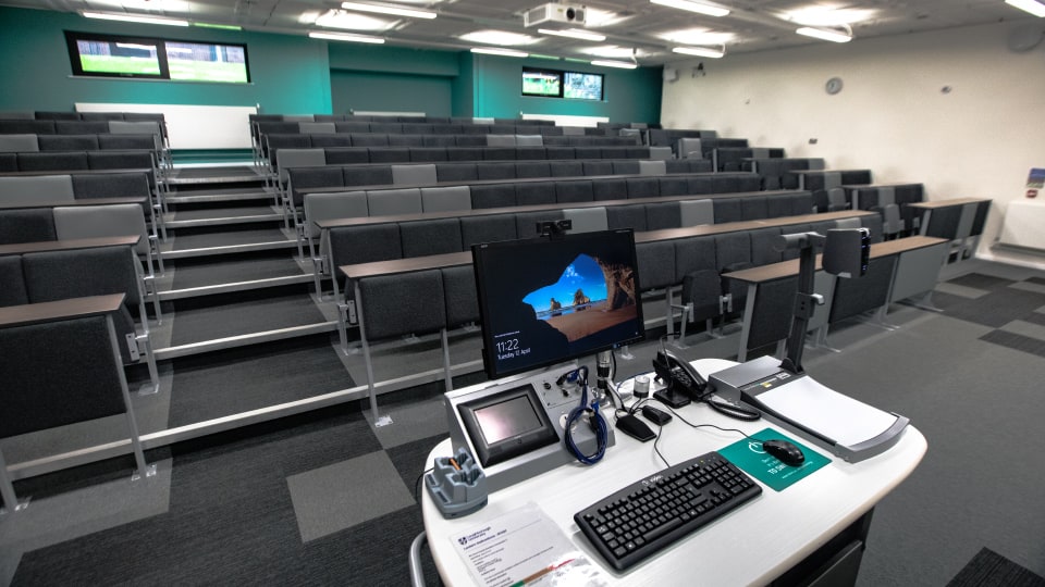 Frank Gibb RT037 - Lecture theatre with tiered seating, a teaching podium featuring a computer, keyboard, mouse, and various electronic devices.