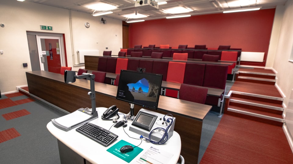 Frank Gibb RT040 - Lecture theatre with tiered seating, red and maroon chairs, a lecturer's desk with computer, keyboard, phone, and projector controls.