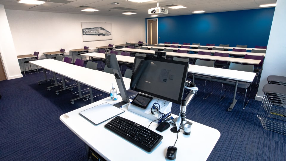 S Building S231 - Teaching room with flat tables, chairs, and a lectern with computer equipment. Blue accent wall and overhead projector.
