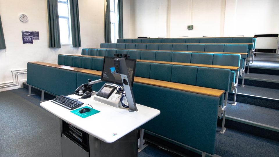 Schofield SCH105 - Lecture theatre with tiered seating, teal upholstery, and a lecturer's podium equipped with a computer, keyboard, and microphone.