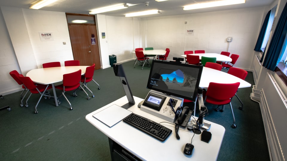 Brockington extension U112 - Small teaching room with flat layout, grouped tables, projector screen at front, and ceiling lighting.