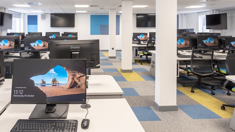 Teaching Room WAV001 - Spacious computer lab with rows of desks, monitors displaying a beach scene, ergonomic chairs, and colourful carpet tiles.