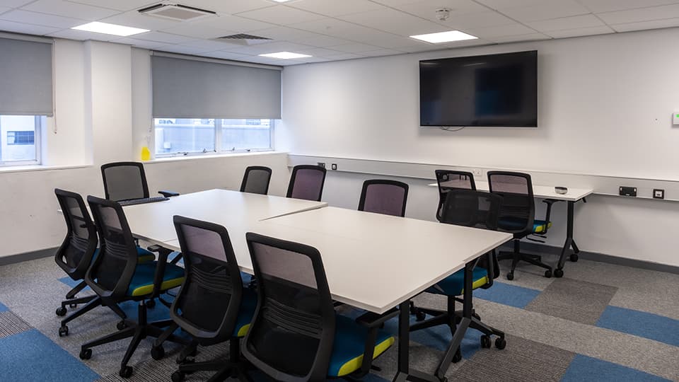 Teaching Room WAV002 - Teaching room with large rectangular table, twelve office chairs, wall-mounted TV, and windows with gray blinds.