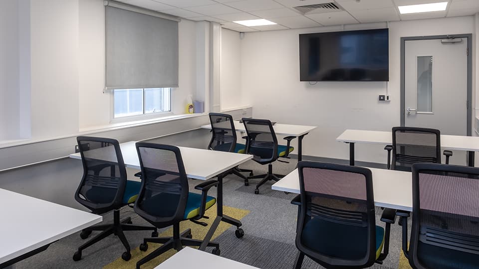 Teaching Room WAV003 - Classroom with white tables, black mesh chairs, large wall-mounted screen, window with blind, and door. Bright, modern design.