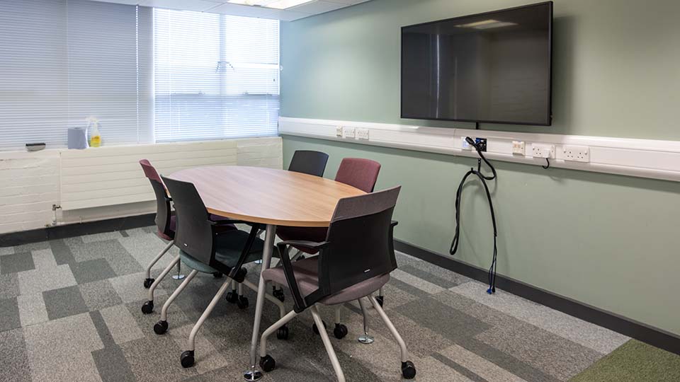 Teaching Room WAV008 - Teaching room with oval table, five chairs, wall-mounted TV, power outlets, and window blinds. Neutral colours, carpeted floor.