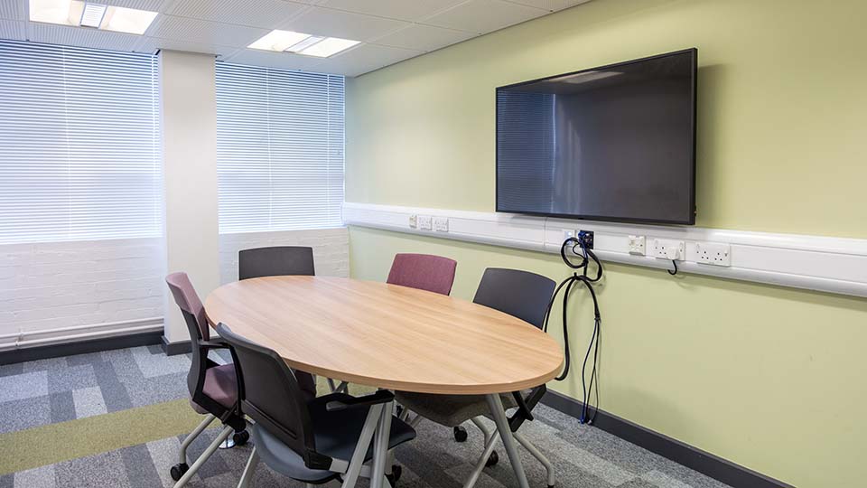 Teaching Room WAV009 - Teaching room with oval table, five chairs, wall-mounted screen, green walls, and large windows with blinds.