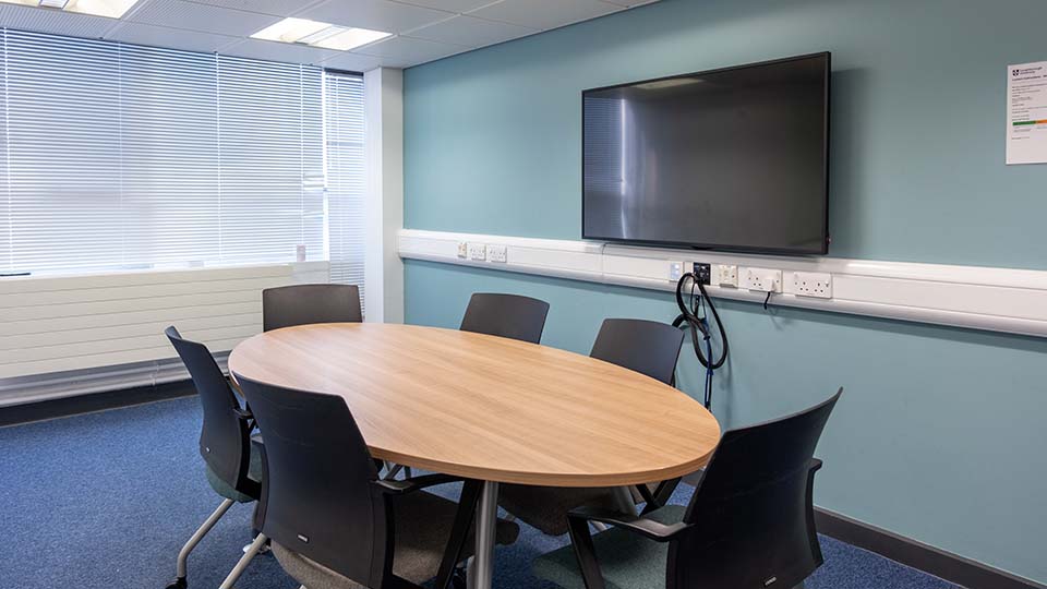 Teaching Room WAV010 - Teaching room with oval table, six chairs, wall-mounted TV, blue walls, and large window with blinds.