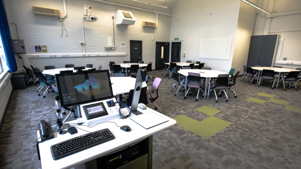 Wavy Top WAV011 - Modern teaching room with circular tables, chairs, and a central instructor's desk featuring a computer and control panel.