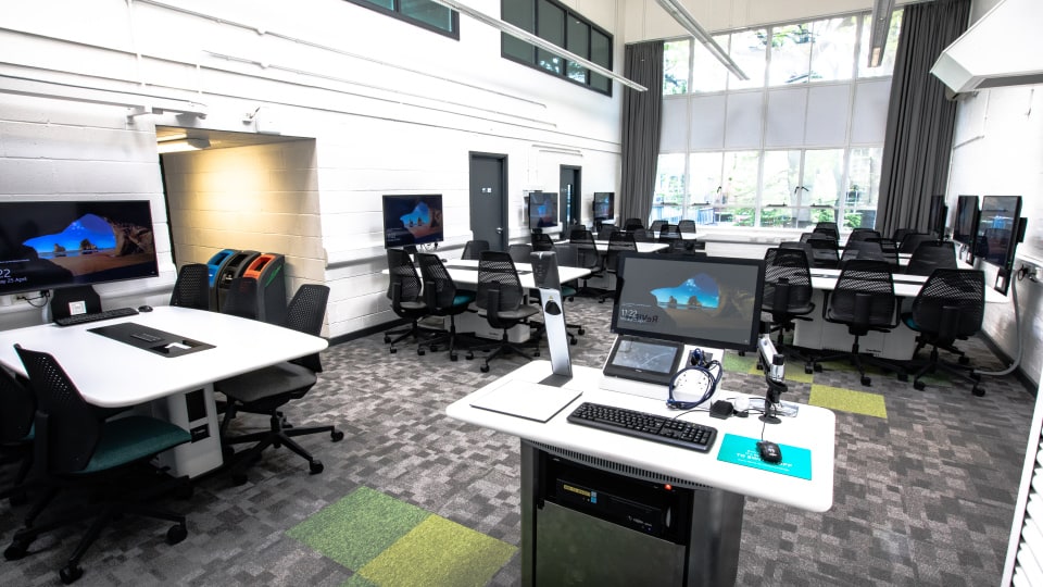Wavy Top WAV017 - Modern computer lab with multiple workstations, swivel chairs, large windows, and a central teaching podium with screens and equipment.