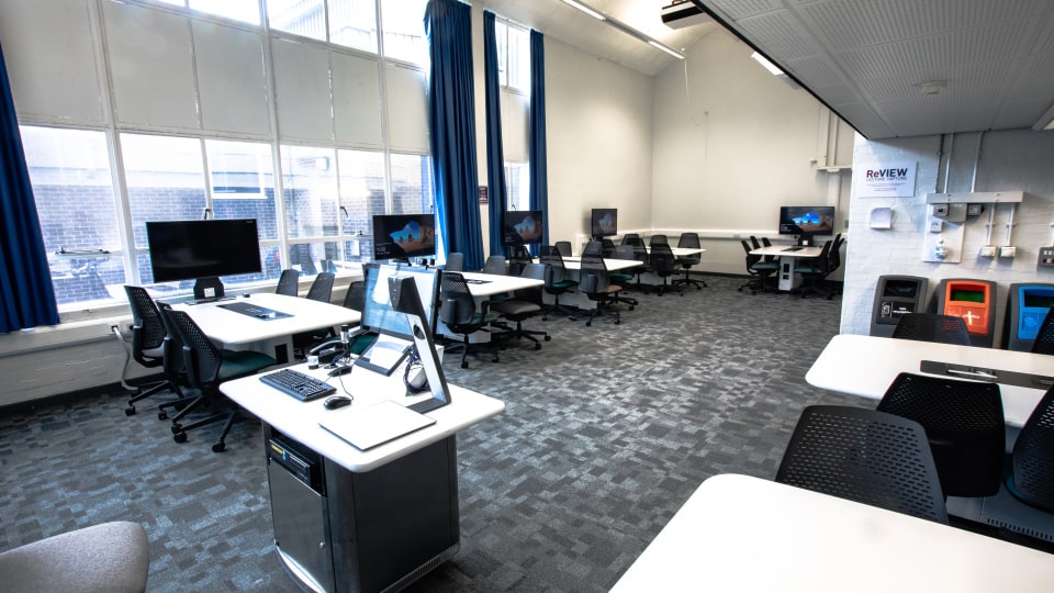 Wavy Top WAV019 - Spacious teaching room with multiple computer workstations, large windows, blue curtains, and a central instructor's desk equipped with technology.