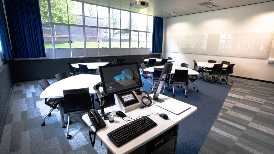 Wavy Top WAV027 - Modern teaching room with hexagonal tables, chairs, large windows, blue curtains, and a presentation desk equipped with computer and phone.