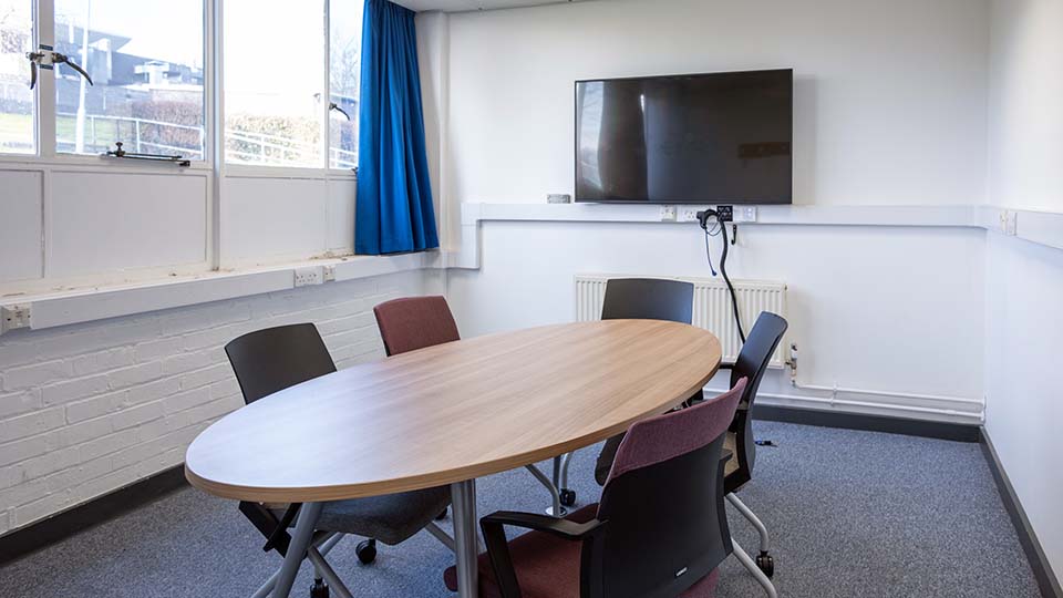 Wavy Top WAV031 - Small teaching room with an oval table, five chairs, wall-mounted TV, large windows, and blue curtain.