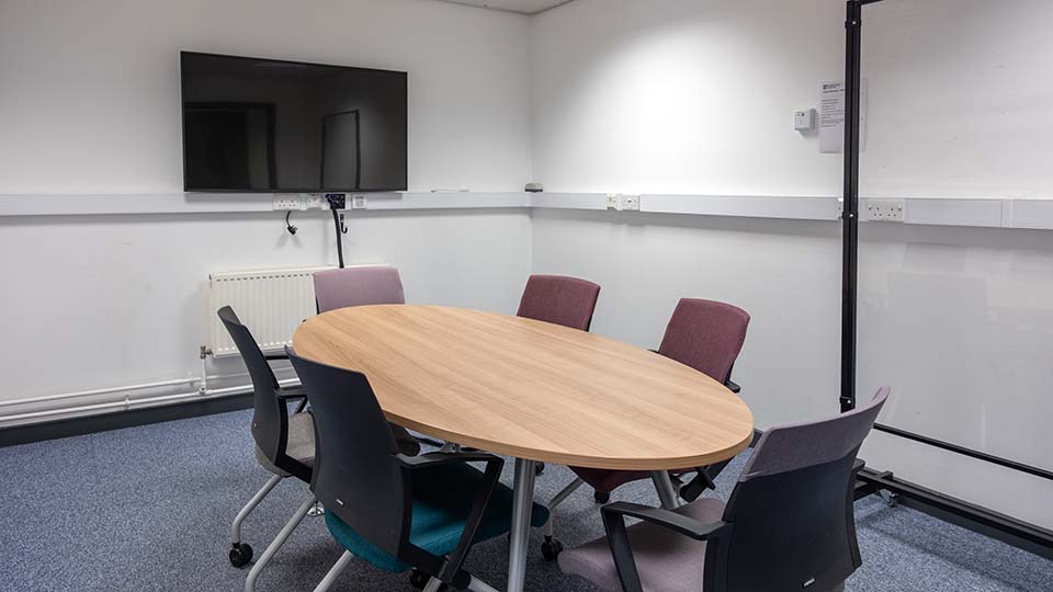 Wavy Top WAV032 - Oval table with six chairs in a teaching room. Wall-mounted TV, whiteboard, and power outlets visible. Neutral decor.