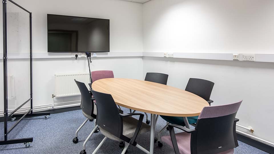 Wavy Top WAV033 - Teaching room with oval table, six chairs, wall-mounted TV, whiteboard on wheels, and power outlets along the wall.