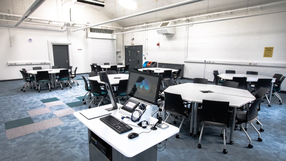 Wavy Top WAV041 - Modern teaching room with hexagonal tables, black chairs, and a central instructor's station featuring a computer and control panel.
