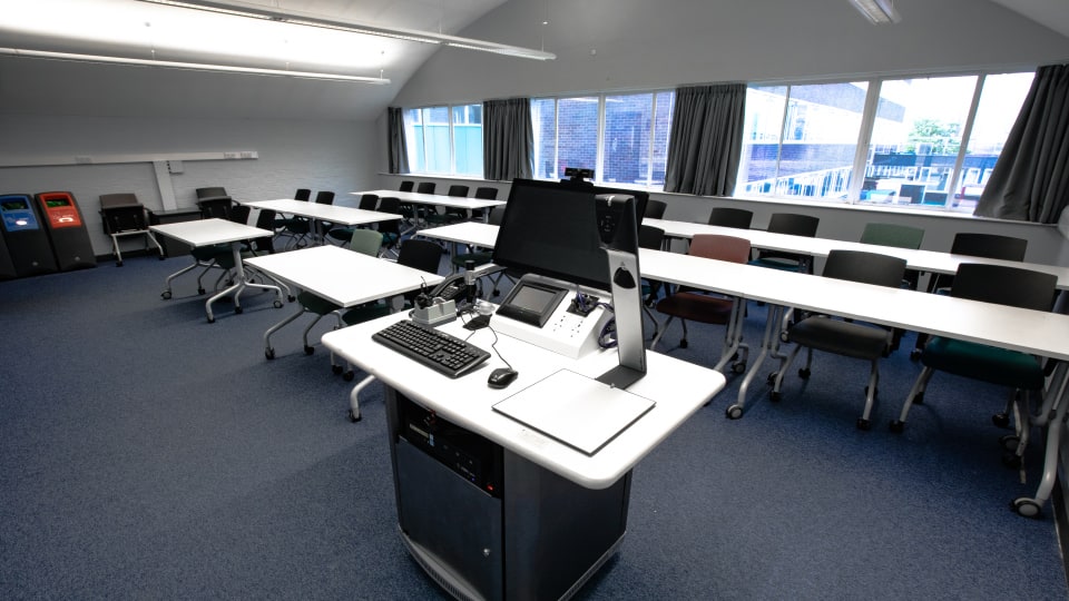 Wavy Top WAV046 - Teaching room with a podium, computer, and microphone. Rows of tables and chairs face large windows with curtains.