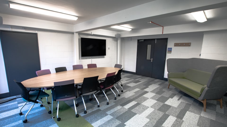 Wavy Top WAV068 - Teaching room with oval table, eight chairs, wall-mounted screen, and a green cushioned sofa. Gray and green patterned carpet.